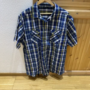 Blå-rutig kortärmad skjorta - Kortärmad skjorta i blått, vitt och gult rutmönster. Skjortan har klassisk krage, två bröstfickor med lock och knappar framtill. Materialet känns lätt och passar bra till varmare dagar.