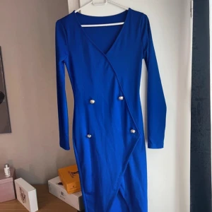 Kornblå omlottklänning med knappar - Snygg kornblå långärmad klänning med omlott-design och dekorativa silverfärgade knappar framtill. Klänningen har v-ringning och figurnära passform, perfekt för dig som vill sticka ut med en färgstark look.