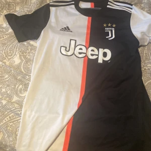 Juventus Adidas fotbollströja svart/vit - Juventus matchtröja från Adidas med klassisk svart och vit design, röd linje i mitten och Jeep-logga på bröstet. Tröjan har korta ärmar, rund hals och tre vita ränder på axlarna. Broderat Juventus-emblem med tre stjärnor. Tillverkad i lätt och ventilerande material.
