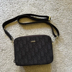 Svart axelväska från Dior - Snygg svart axelväska från Dior med diskret mönster och silverfärgad logga framtill. Väskan har justerbar axelrem och dragkedja upptill. Perfekt storlek för det viktigaste och en stilren accessoar till din outfit.