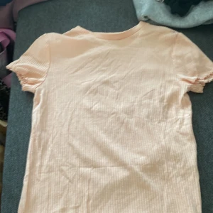 Ljusrosa ribbad topp från H&M - Supersöt ljusrosa ribbad topp från H&M med korta ärmar och volangdetalj vid ärmslut. T-shirten har en slim passform och är tillverkad i mjuk bomull, perfekt för en chill och trendig look.