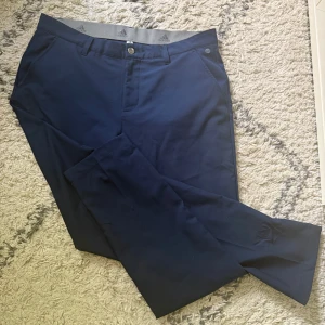 Mörkblå golfbyxor från Adidas - Snygga mörkblå golfbyxor från Adidas med klassisk passform och raka ben. Byxorna har diskreta fickor fram och bak samt bälteshällor. Tillverkade i ett mjukt och slitstarkt bomullsmaterial med en stilren look och Adidas-logga på insidan och bak.