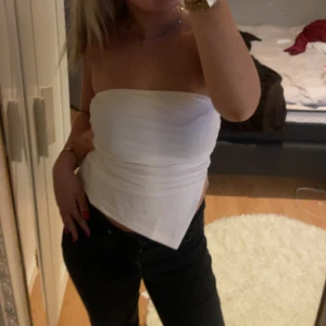 Vit bandeau tubtopp - Säljer en stilren vit bandeau tubtopp med asymmetrisk nederkant. Toppen är ärmlös och sitter tight mot kroppen, perfekt att matcha med jeans eller kjol. Enkel och fräsch look som passar till många olika stilar.