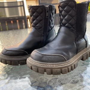 Svarta quiltade boots med dragkedja - Tuffa svarta boots med grov sula och quiltad detalj på skaftet. Skorna har dragkedja på sidan för enkel påtagning och är tillverkade i skinnliknande material. Perfekta för dig som gillar chunky stil och vill sticka ut lite extra.