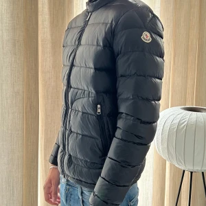 Svart Moncler dunjacka  - Säljer en svart dunjacka från Moncler med klassisk quiltad design och hög krage. Jackan har dragkedja framtill, två fickor med dragkedja och Moncler-logga på ärmen. Perfekt för kyliga dagar och riktigt snygg till jeans eller cargo.