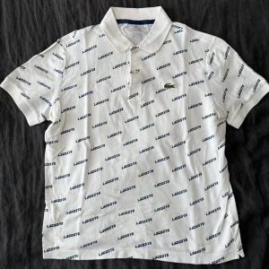 Vit pikétröja Lacoste - Snygg vit pikétröja från Lacoste i storlek S. Tröjan har korta ärmar, klassisk krage och knäppning framtill. All-over print med Lacoste-loggan i mörkblått och den ikoniska gröna krokodilen broderad på bröstet. Perfekt för en clean och sportig look.
