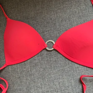 Röd bikini från Calvin Klein - Snygg röd bikini från Calvin Klein med trekantsformade kupor och en silverfärgad ring med logga i mitten. Lappen med storleken är borta men kan säkert kolla på något sätt. Bikinin har tunna knytband och är perfekt för stranden. Materialet känns mjukt och stretchigt, troligtvis syntet.