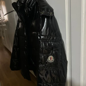 Svart Moncler pufferjacka med logga - Säljer en svart, glansig pufferjacka från Moncler med klassisk logga på ärmen. Jackan har dragkedja framtill, hög krage och praktiska fickor. Perfekt för dig som vill ha en stilren och varm jacka med exklusiv känsla.