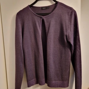 Lila kofta från Lindex, storlek 38/40 - Lila kofta från Lindex i storlek 38/40. Den är stickad, har rund halsringning och långa ärmar. Koftan är öppen framtill utan knappar och har ribbade muddar med lite glitter vid ärmslut och nederkant.