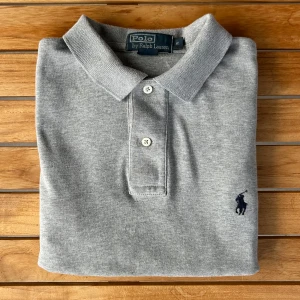 Ralph Lauren Piké Grå S - Klassisk grå pikétröja från Polo Ralph Lauren i 100% bomull. Tröjan har korta ärmar, krage med två knappar och den ikoniska broderade loggan på bröstet. Perfekt basic-plagg med snygg passform och stilren look.