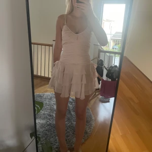 Ljusrosa volangklänning med tunna axelband - Supersöt ljusrosa kort klänning med tunna axelband och v-ringning. Klänningen har rynkad överdel och flera lager volanger i kjolen som ger en flowy känsla. Perfekt för sommarens alla tillfällen och ger en romantisk vibe. Är omsydd i midjan för en mer snatch känsla 