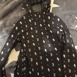 Juventus vindjacka Adidas XS - Svart vindjacka från Adidas med Juventus-logga, vita stjärnor och hästar över hela jackan. Jackan har huva, hel dragkedja och är tillverkad i lätt polyester. Perfekt för dig som älskar fotboll och vill sticka ut med en sportig look.