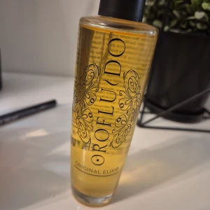 Orofluido Original Elixir olja - Orofluido Original Elixir är en lyxig hårolja i glasflaska med svart kork. Oljan är gyllengul och har en elegant design med svarta mönster på flaskan. Perfekt för att ge glans och mjukhet till håret.