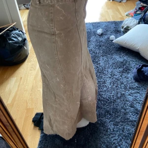 Beige lång jeanskjol med sömdetaljer - Säljer en beige långkjol i jeansmaterial med coola sömdetaljer och hög midja. Kjolen har knapp och dragkedja framtill samt bälteshällor. Snygg, figurnära passform upptill och lite utsvängd nertill för en trendig siluett.