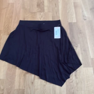 Svart asymmetrisk kjol från Nelly - Svart kortkjol från Nelly i modellen Lovely Asymmetric Skirt. Kjolen har en snygg asymmetrisk nederkant som ger en cool och modern vibe. Tillverkad i mjukt och lätt material, perfekt för dig som gillar stilrena plagg med en twist.