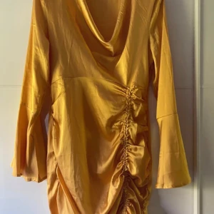 Gul satin klänning med drapering - Snygg gul kortklänning i satin med långärmade utsvängda ärmar. Klänningen har en draperad cowlneck-ringning och cool rynkdetalj längs ena sidan framtill som ger en unik look. Perfekt för dig som vill sticka ut med en trendig och färgstark outfit. Man kan justera längden på klänningen med banden. Använd en gång.