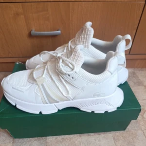 Vita sneakers från Lacoste - Snygga vita sneakers från Lacoste med chunky sula och coola detaljer. Ovandelen är i mesh och syntet med mönstrad plös och tydlig Lacoste-logga. Skorna har snörning och sportig vibe, perfekta för dig som gillar stilrena och fräscha sneakers. Box följer med 