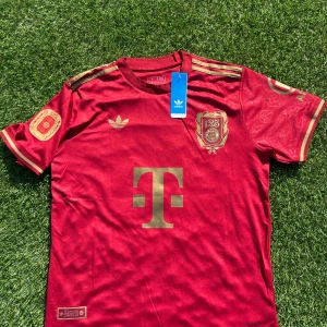 Bayern München Müller 25 Adidas tröja - Riktigt snygg Bayern München fotbollströja från Adidas i röd med guldiga detaljer och tryck. Müller 25 på ryggen, klubbmärke och sponsorlogga framtill. Korta ärmar, slim fit och tillverkad i lätt, ventilerande material. Perfekt för dig som älskar fotboll och vill sticka ut.