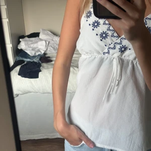 Vit gullig topp med blåa detaljer  - Supersöt vit topp med tunna axelband och v-ringning. Ovanför bysten finns blå broderade blommor och dekorativa detaljer. Den är luftig och skön vilket är perfekt nu i sommar☀️den är från shein och är i storlek xs. Vad jag kommer ihåg så är den inte använd 