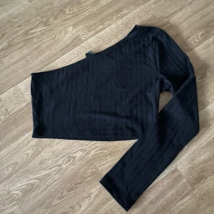 Svart oneshoulder croppad topp - Snygg svart croppad topp med oneshoulder-design och lång ärm. Tunt ribbat tyg som ger en stilren look. Perfekt för dig som vill sticka ut med en asymmetrisk stil.