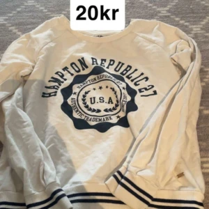 Vit sweatshirt från Hampton Republic - Vit långärmad sweatshirt från Hampton Republic med marinblå tryck och detaljer. Tröjan har rund halsringning och ribbade muddar med blå ränder vid ärmslut och nederkant. Perfekt för en sportig och avslappnad stil.
