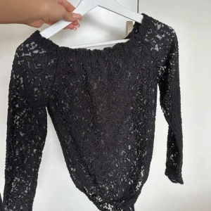 Svart spetsblus med lång ärm - Supersnygg svart blus i spets med lång ärm och offshoulder-skärning. Toppens genombrutna spetsmönster ger en cool och elegant vibe. Perfekt att styla med jeans eller kjol för en trendig look.
