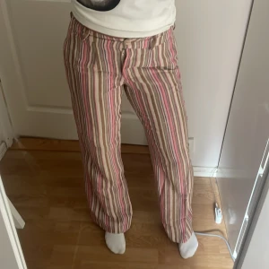 Randiga linnebyxor från Flare, strl 36 - Snygga linnebyxor från Flare i storlek 36 med vertikala ränder i rosa, beige, brunt och vitt. Byxorna har normal passform, raka ben och klassisk midja. Perfekta för en chill och trendig look under varmare dagar.