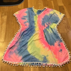 Färgglad tie-dye kortklänning med bollar - Superfin kortklänning i tie-dye med blått, rosa och gult. Klänningen har v-ringning och ärmlös design, samt vita bollar längs kanterna för en extra söt touch. Perfekt för sommarens varma dagar och strandhäng.