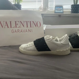 Valentino Garavani skor - Snygga vita Valentino Garavani Rockstud sneakers med svart band och ikoniska nitar. Skorna har rund tå, snörning och platt sula. Tillverkade i skinn för en lyxig känsla och stilren look. Perfekta för dig som vill sticka ut med exklusiva detaljer. Skriv innan köp och skriv vud funderingar