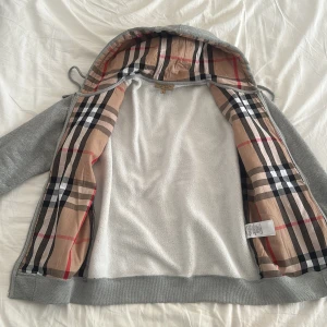 Grå hoodiejacka från Burberry M - Grå hoodiejacka från Burberry i bomull med dragkedja och klassiskt rutigt foder i beige, svart och rött. Jackan har broderad logga på bröstet och mjukt innerfoder. Perfekt för dig som vill ha en stilren och lyxig look med ikoniska Burberry-detaljer. 