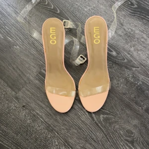 EGO klackar med transparent rem - Snygga klackar från EGO med transparenta remmar och klack i plexiglas. Skon har en nude/ljusbeige bas och öppen tå. Perfekt för dig som vill ha en clean och modern look med en twist.