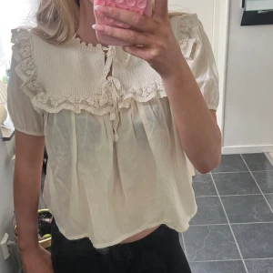 Söt vit blus med spets och volang - Gullig vit blus med korta puffärmar, virkade spetsdetaljer och volang längs axlar och bröst.. Perfekt för en gullig look. Har dock en sminkfläck på insidan av blusen❤️