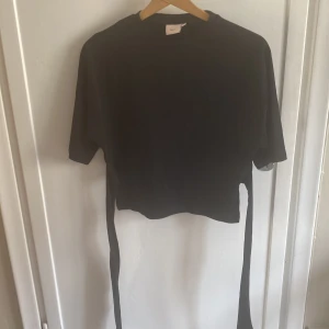 Svart croppad topp med knyt från Nelly - Svart croppad topp från Nelly med korta ärmar och långa knytband i midjan som kan knytas i ryggen. Enkel och stilren design, perfekt att matcha med höga jeans eller kjol. Mjuk och bekväm bomullskänsla. Andvänd en gång och är skit snygg