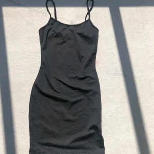 Svart tight kortklänning med smala band - Enkel och stilren svart kortklänning med smala axelband och figurnära passform. Klänningen har en rak halsringning och är ärmlös, perfekt för en clean look. Materialet känns mjukt och stretchigt, vilket gör den bekväm att bära hela dagen.