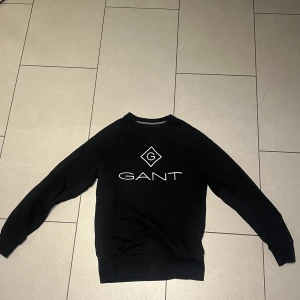 Svart sweatshirt från GANT XS - Svart sweatshirt från GANT i storlek XS med klassisk logga framtill. Tröjan har rund halsringning, långa ärmar och ribbade muddar vid ärmslut och nederkant. Perfekt för en clean och stilren look.
