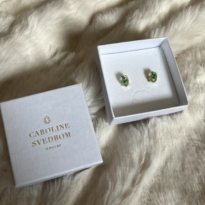 Gröna örhängen från Caroline Svedbom - Snygga örhängen från Caroline Svedbom med droppformade gröna kristaller infattade i guldfärgad metall. Perfekta för att ge din look lite extra bling och färg. Kommer i originalask. Nypris 375kr. 🫶🏻