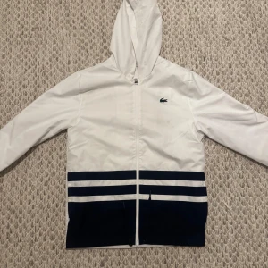 Lacoste windbreaker - Snygg vit vindjacka från Lacoste med marinblå detaljer och två breda ränder längst ner. Jackan har huva, dragkedja och stor Lacoste-logga på ryggen. Perfekt för dig som gillar sportig och clean stil.