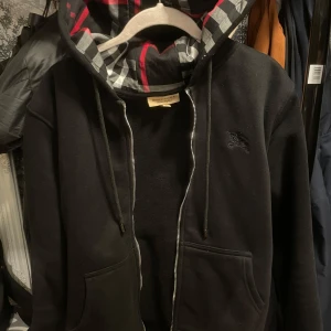 Svart Burberry hoodie  - Svart hoodie från Burberry med dragkedja och klassiskt rutigt foder i huvan i svart, vitt och rött. Tröjan har snörning vid huvan, två fickor framtill och diskret broderad logga på bröstet. Perfekt för dig som gillar stilrena och lyxiga streetwear-plagg.