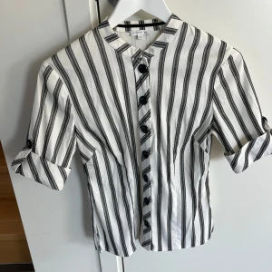 Randig skjorta från STOCKH LM - Snygg skjorta från STOCKH LM i vitt med svarta vertikala ränder. Skjortan har svarta knappar framtill, ståkrage och uppvikbara ärmar med knapp. Tillverkad i bomull för en skön känsla och stilren look.