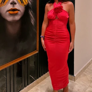 Rödrosa halterneck långklänning med ros - Säljer en snygg röd långklänning med halterneck och en stor rosdetalj vid halsen. Klänningen har åtsittande passform och rynkade detaljer framtill som framhäver figuren. Perfekt för dig som vill sticka ut med en elegant och trendig look. Använd 2 gånger.