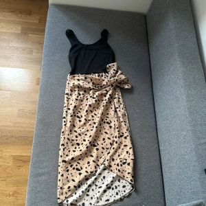 Sommarklänning  - Supersnygg klänning med svart ribbad överdel och omlottkjol i beige med svarta leopardliknande prickar. Kjolen har en stor rosett i midjan och är längre baktill. 