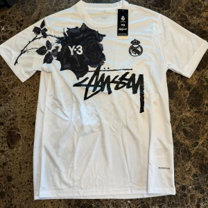 Vit stussy t-shirt M - Snygg vit t-shirt från Y-3 x Real Madrid med svart ros-tryck både fram och bak, samt cool Stüssy-liknande logga på bröstet. Tillverkad i luftig polyester med Aeroready-teknik. Perfekt för dig som gillar streetwear och fotbollsvibe.