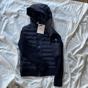 Mörkblå Moncler cardigan med huva(går inte att ta bort huva) - Snygg mörkblå pufferjacka från Moncler med stickade ärmar och quiltad kropp. Jackan har huva, dragkedja framtill och en liten ficka med dragkedja på ärmen. Perfekt för vindigare dagar och riktigt stilren look.