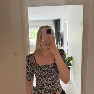 Svart blommig topp från Lindex XS - Supersöt svart topp från Lindex i storlek XS med vita småblommor över hela. Toppen har fyrkantig ringning, puffiga axlar och trekvartsärm. Den är figurnära och ribbad i materialet, vilket gör den både bekväm och snygg.