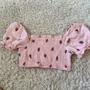 Rosa croppad blus med jordgubbar - Supersöt rosa croppad blus från Cotton On med puffiga korta ärmar och broderade jordgubbar över hela toppen. Smockad överdel och volangdetaljer vid ärmslut och halsringning. Perfekt för dig som vill sticka ut med en lekfull och somrig vibe.