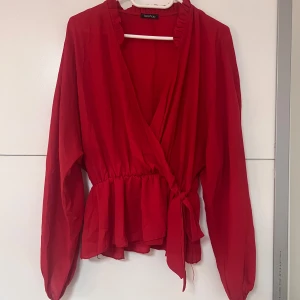 Röd omlottblus från Boohoo -  Röd omlottblus från Boohoo med långa ärmar och volangdetaljer vid kragen. Blusen har knyt i midjan och resår vid ärmsluten för en snygg passform.
