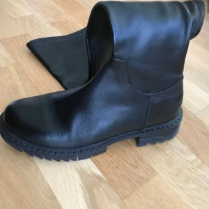 Svarta höga skinnstövlar med dragkedja - Snygga svarta stövlar i skinn med chunky sula och grovt mönstrad yttersula. Modellen är knähög med dragkedja på insidan för enkel påtagning. Perfekta för att lyfta din outfit med en edgy vibe.