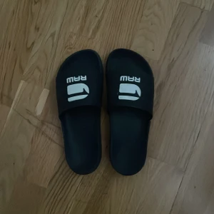 Gstar raw tofflor - Svarta sandaler från RAW med vit logga på ovandelen. Klassisk slip-on modell med bred rem över foten och platt sula. Perfekta för sommaren eller att chilla hemma. Tillverkade i syntetmaterial för enkel rengöring