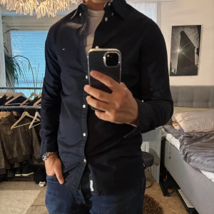 Mörkblå skjorta Tommy Hilfiger slim fit - Snygg mörkblå skjorta från Tommy Hilfiger i slim fit-modell. Skjortan har klassisk krage med knappar, vita knappar framtill och diskret logga på bröstet. Tillverkad i mjuk bomull och passar perfekt till jeans eller chinos.
