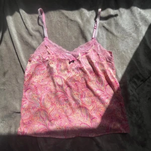 Paisleymönstrad rosa linnetopp med spets - Supersöt linnetopp i rosa och lila paisleymönster med tunna axelband. Toppen har spetskant upptill och en liten rosett med hjärtberlock framtill. Materialet känns mjukt och svalt, perfekt för varma dagar. 